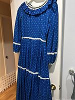 Full view of blue polka-dot pioneer dress hanging, showing lace-trimmed ruffled collar, long sleeves, tiered skirt
