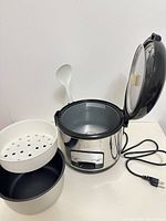 Rice cooker with lid open, inner pot, steamer basket, spatula and power cord visible