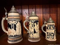 Front view of the three steins showing relief scenes and lids
