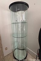 Full view of cylindrical glass cabinet showing three shelves, black top and base