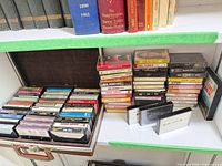 Shelf view showing brown carry case open with rows of cassette tapes and additional stacks of tapes