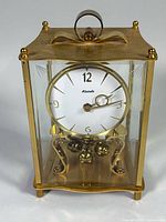Front angled view of brass and glass anniversary clock showing case, dial, pendulum