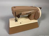 Left front view of Singer Sewhandy toy sewing machine showing tan and cream color, crank handle, base