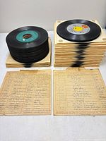 Two stacked bundles of approximately fifty 45 RPM records in tan sleeves beside two handwritten index sheets