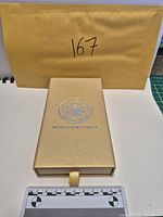 Closed gold-tone presentation box with ruler for scale