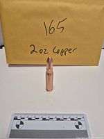 Front view of copper bullet-shaped bar standing upright beside envelope labeled 165 and ruler