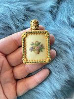 Front view of gold metal perfume pendant showing needlepoint floral panel and green rhinestone border