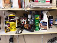 Full view of shelf with assorted cleaners, sprays, Bona bottles, Raid can, butane torch