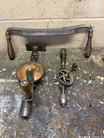 Three tools on workbench: drawknife at top, two hand drills below