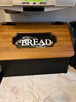 Top view of bread box showing wood-grain lid, glass window with “BREAD” text, and hinges