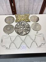 Full lot showing seven metal pieces: four round pierced trivets/coasters, one round flatware-motif trivet, one brass butterfly trivet, one expandable rack