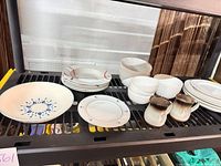 Shelf with assorted dinnerware items including blue floral plate, stack of red-accent plates, plain white bowls and two creamers