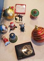 Assorted ornaments laid out: gold ball, moose, Santa, donkey, red-green top, porcelain bell, snow globe, large red striped ball, boxed gold ornament