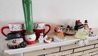 Full lot on mantle showing mugs, candle holder, tall hat snowman, assorted figurines and snow globes