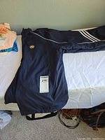 Full view of navy Adidas Notre Dame windbreaker jacket on bed showing ND crest, white shoulder piping, sleeve stripes, zipper front