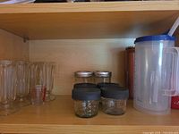 Shelf showing four footed sundae glasses, measuring shot glass, four small jars with black lids, two mason jars with metal lids, copper-tone tumbler and plastic pitcher with blue lid