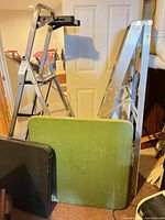 Front view showing silver aluminum ladder, black-tread step ladder, green folding table, edge of black table