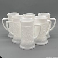 Front angled view of six white milk glass tumblers with embossed scenes