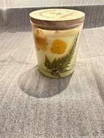 Front view of candle showing pressed flowers and ferns on yellow wax in glass jar with wood lid