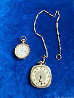 Two pocket watches, gold-tone round Elgin and silver-tone cushion Birks with chain, on blue background