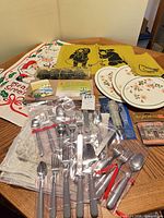 Group shot of flatware in bags, floral trivets, boxed coasters, cedar plank, yellow tea towels, Christmas tea towel, casserole stand partially visible