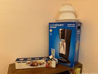 SodaStream Genesis boxed unit, Black & Decker EK300 knife box, three white jelly molds stacked on top, two white plastic accessories in front