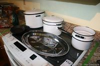 Four white enamelware pots with lids, including a double boiler and a canning pot, displayed on a stove along with a stainless steel meat serving tray with handles.