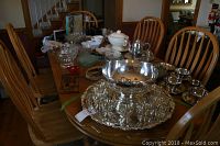 Oak pedestal dining table with assorted glass, ceramic, and silverware items arranged on top, shown with wooden chairs around.