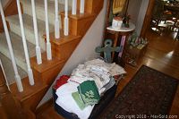 Photo showing assorted linens including embroidered tablecloths and napkins stacked beside a wooden stair rail with a green painted wooden anchor wall decor visible.