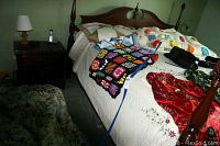 Overview of queen bed with white hand crocheted bedspread, assorted pillows, multicolored afghan, king size floral comforter on floor, red silky fabric and black hat on bed.