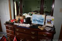 View of ladies' wares on top of wooden dresser with mirror, including painting, purses, gloves, and latch hook kit.