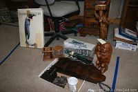Photo of an assortment of decorative and collectible items on carpeted floor with carpet tape markings. Items include a framed wall hanging of Emperor Penguin, wooden carved figurines, an inflatable penguin, a locomotive phone, wooden bookends, a white mug, glass jar, and pipes on wooden stand.
