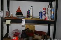 Shelf with assorted hand tools and garage chemicals including red drill bit case, palm sander, various bottles and tubes