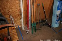 Several long-handled yard tools including shovels, post hole digger, post digger tool, and a green handle tool arranged against shed wall.