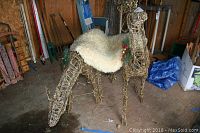 Two woven twig deer lawn decorations with light strings, one with antlers, sitting in a wood shed surrounded by various tools and equipment.