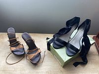 Both pairs of shoes on tabletop: tan lace-up heels left, black ribbon heels on shoebox right