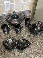 Entire Mikasa black floral porcelain tea set displayed on countertop: teapot, two upright cups, stack of additional cups nested on saucers, and divided leaf dish