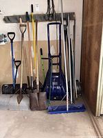Group of 10+ assorted shovels, rake, hoe, poles leaning against garage wall
