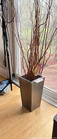 Silver-gray rectangular metal vase with brown twig branches and faux calla lily shown beside window