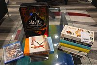 Overall lot on glass table: Buffy Season 2 tin, Battlestar Galactica Blu-ray, fitness DVDs stack, CDs stack, paperbacks stack