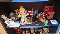 Shelf showing multiple Disney figurines, doll, plush, ornaments and stands