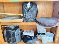 Shelf showing Ninja multi-cooker, Instant Pot booklet, black lid, placemats, linens, boxed waffle maker and bag