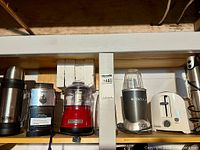 Shelf showing six kitchen appliances: Thermos, Capresso grinder, KitchenAid chopper, NutriBullet blender, Proctor Silex toaster, immersion blender