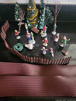 All miniature Christmas village accessories laid out: figures, trees, fences, brick sheet, wreath, bench, snow mound, teddy bear