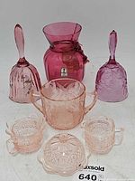 Group shot of all seven pink glass items: cranberry vase, two bells, poinsettia bowl, Moser miniature creamer, sugar bowl and lid