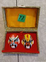 Open display box showing two colorful miniature Chinese opera masks on red velvet base, yellow lining, lot sticker 77 visible