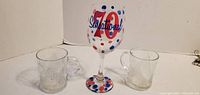 Front view of painted wine goblet centered between two clear glass mugs