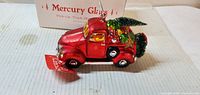 Side view of red mercury glass pickup truck ornament with attached tag and box in background