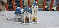 Front view of ceramic village house showing blue roof, wreath-adorned doors, steeple, and cord