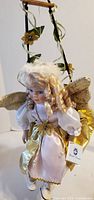 Front view of porcelain angel doll seated on swing showing dress, wings and hang tag